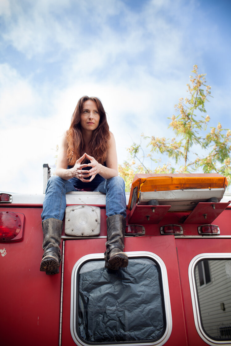 Eliza Jane Schneider sits atop a red bus, her jean clad legs dangling over the edge