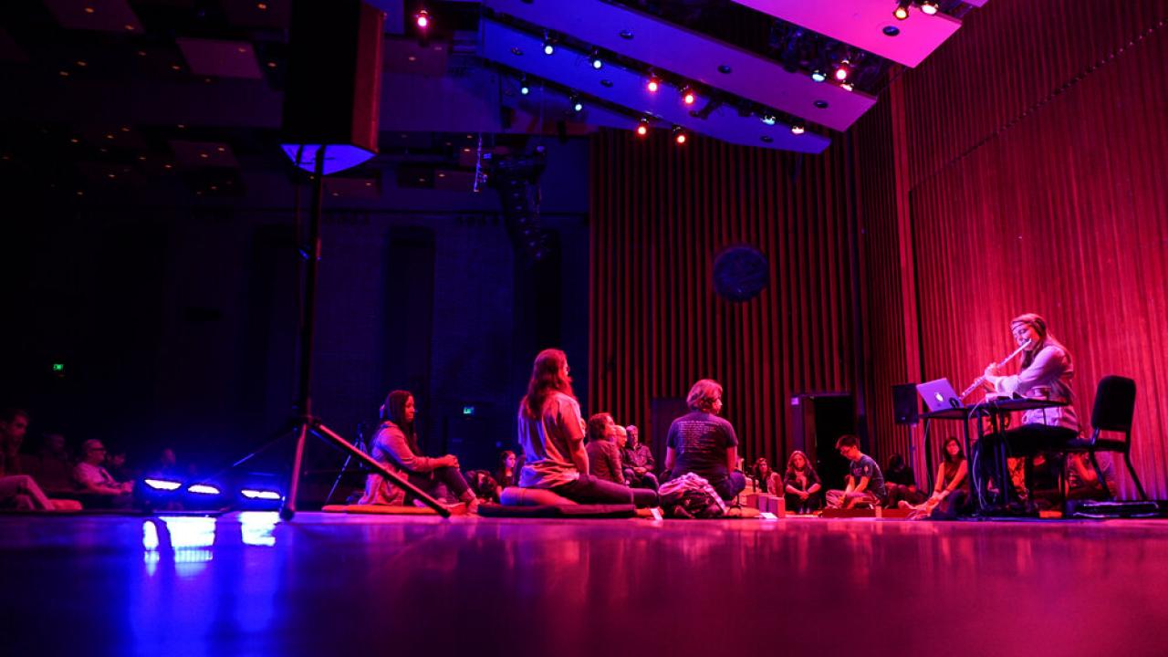 Performer Grace Leslie sits on the right, playing a flute. Audience members sit on the floor in front of her.