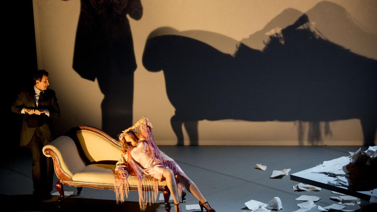 A woman reclines on a chaise lounge, with a man standing beside her. Both cast large shadows behind them