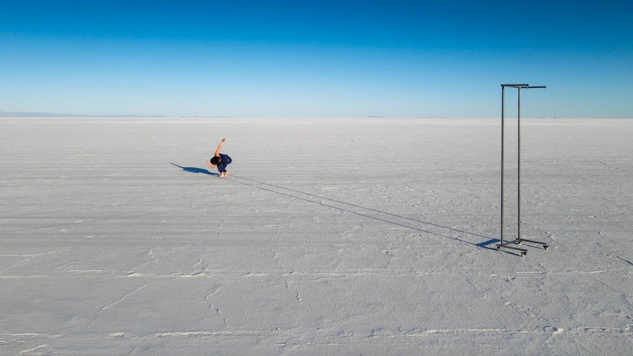The photo is taken outside, with a vast horizon in the background. The ground is snowy, there is a empty rolling coatrack off to the right side. In the center, but back a bit, a dancer is bent over with back arm high and front arm curved.