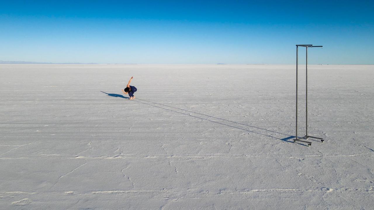 The photo is taken outside, with a vast horizon in the background. The ground is snowy, there is a empty rolling coatrack off to the right side. In the center, but back a bit, a dancer is bent over with back arm high and front arm curved.